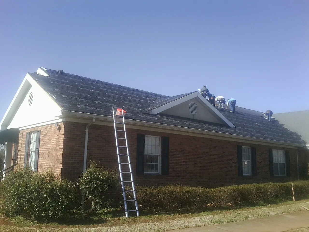Crew tearing shingles off a brick ranch roof with a ladder set — Metal Roof Repair work by our crew in Henniker