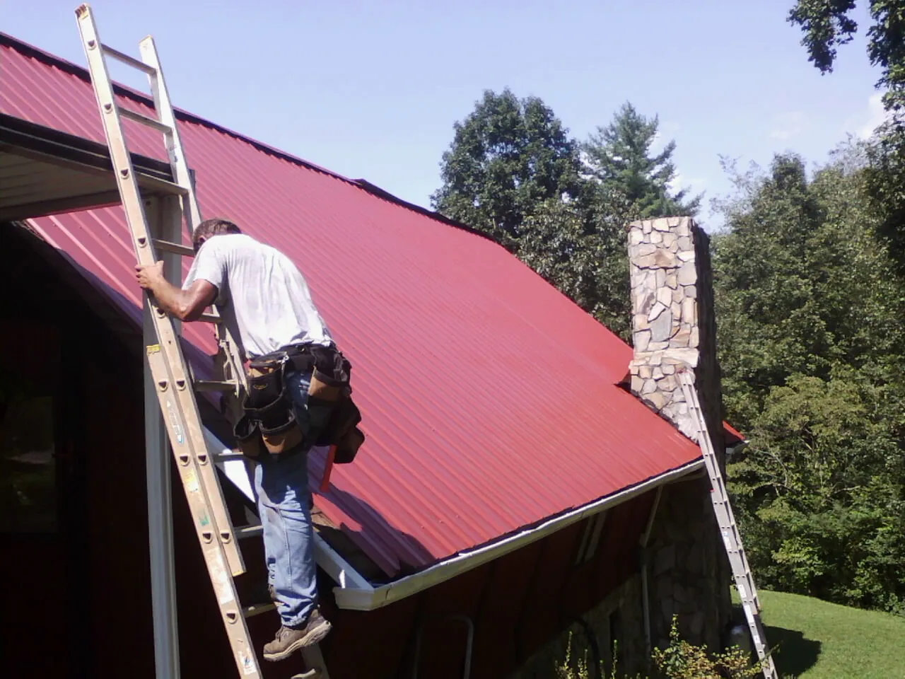Roofer on a ladder beside a red metal roof and stone chimney — Soffit & Fascia Repair by Roofing Brothers in Henniker